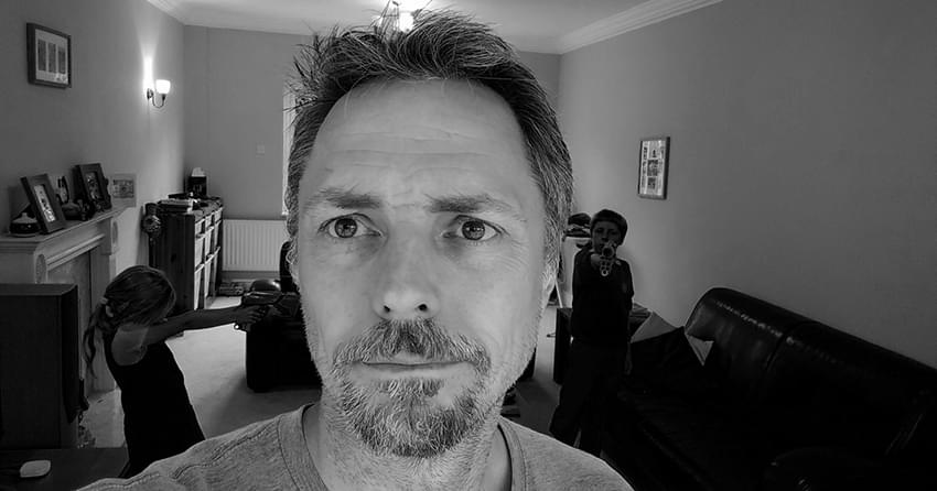 A black and white selfie taken in my lounge, in the background are my kids, both playing with nurf guns.