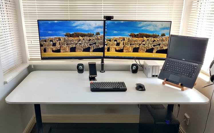 A photograph of a large white computer desk, that includes a keyboard, mouse, mobile phone stand, laptop and stand, and two large monitors.