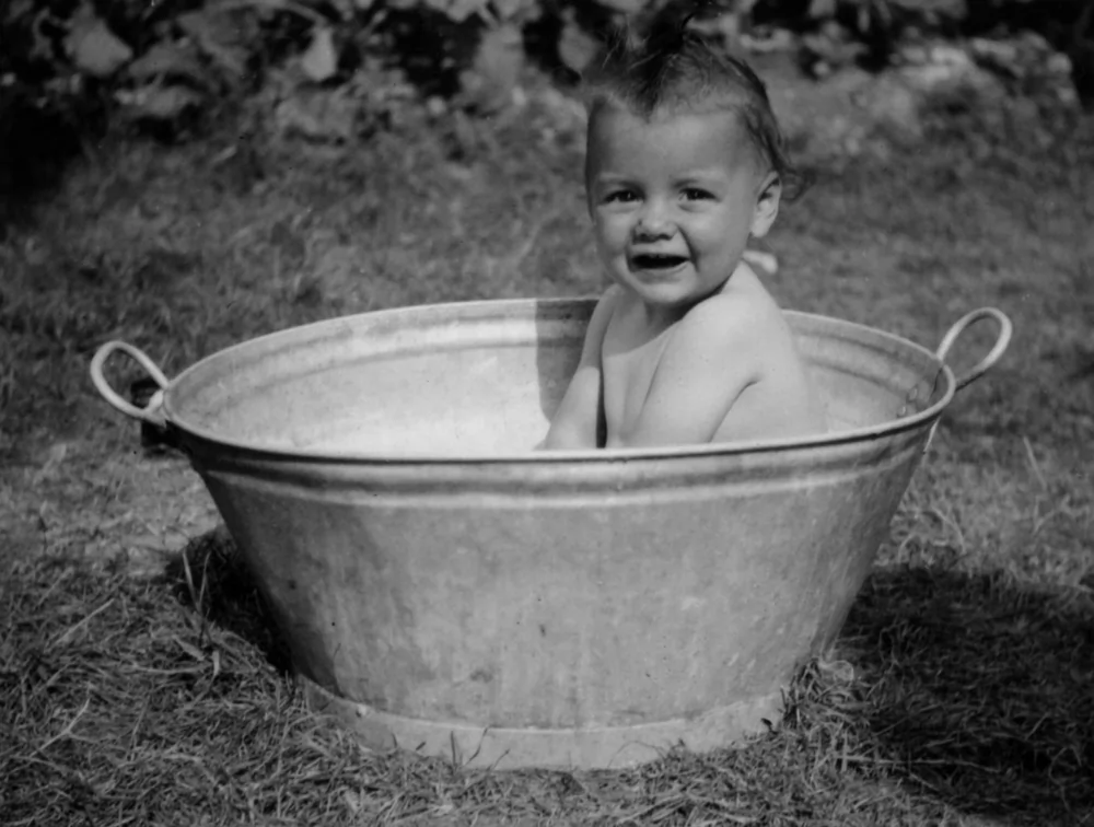 A black and white photograph of a baby in a bathtub located in a garden. Also shown is the full colour restored version.