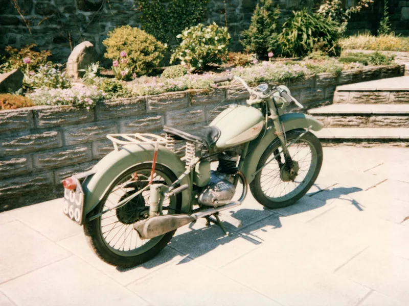 A vintage green motorcycle parked outdoors in front of a tiered stone wall and garden. The second image shows a highly detailed model version of the same motorcycle on a desk, with a box and computer screen displaying its digital design in the background.