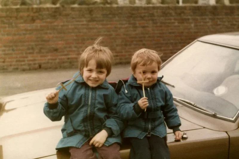 Two young children sit on the bonnet of a car, both wearing blue jackets. In the first image, they are holding crosses, while in the second AI-generated version they are holding ice creams.