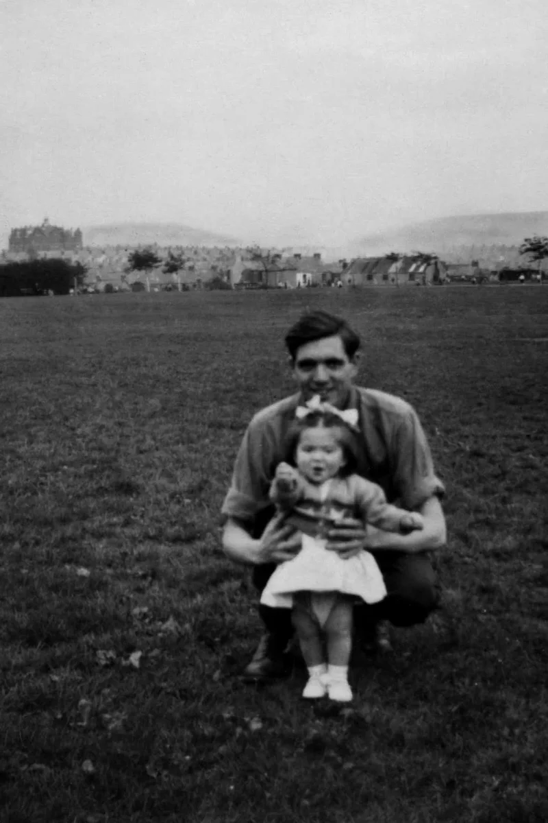 A man crouching on a grassy field holding a young girl standing in front of him, with a backdrop of houses and hills in the distance. The first image shows this scene in black-and-white, while the second is an AI-enhanced color version, bringing vibrant life to the man’s blue shirt and the green grass.