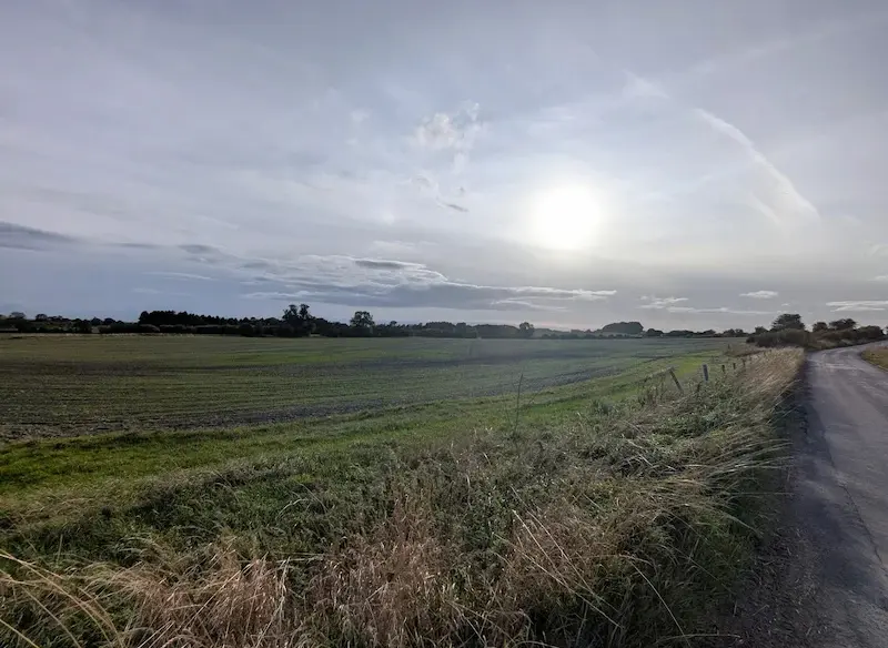 A realistic photograph of a countryside scene with grassy field, a dirt road curving to the right, and a sun shining through a partly cloudy sky. The second image is a vibrant impressionist painting of a rural landscape with a bright sun and dynamic swirling clouds in the sky, showing lush green fields and a dirt path on the right.