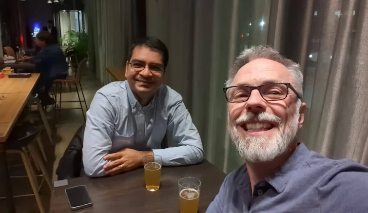 Two men smiling and sitting together at a table with glasses of beer in a relaxed indoor setting. In the next image, a third man, resembling the King of England, has joined them at the table, and all three are smiling for the photo.
