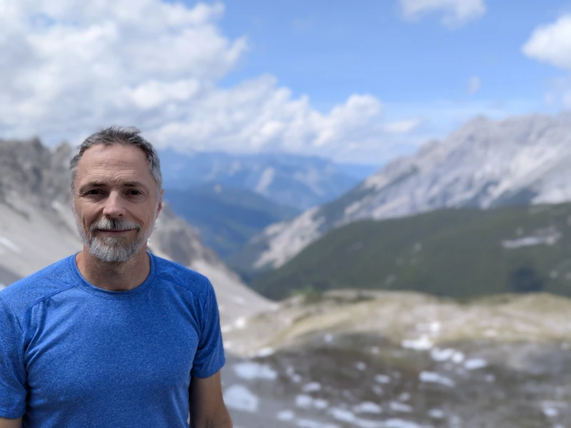 A man wearing a blue t-shirt stands outdoors in front of a mountain landscape under a bright sky in the first image. In the second AI-generated image, the man is depicted as a realistic figurine next to a illustrated box, with a computer screen showing his model in the background.