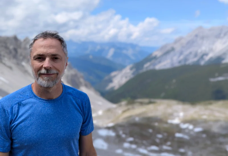 Man with short gray hair and beard standing outdoors in a blue shirt, with a mountainous landscape in the background; default view is a clear, bright photograph and toggles to a dramatic, dark oil painting style of the same scene.
