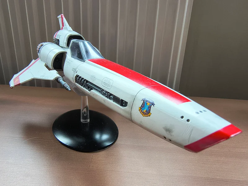 A detailed model of a white and red spaceship (colonial Viper Mark 2 from Battlestar Galactica), shown first on a stand indoors and then in an AI-generated version flying through outer space with a galaxy and stars in the background.