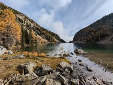 A wide-angle photograph taken from a low vantage point on the bank of a lake. The lake is surrounded by steep mountains on either side. The mountains are covered with pine trees.