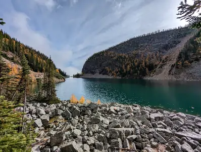 A wide-angle photograph taken from a low vantage point on the bank of a lake. The lake is surrounded by steep mountains on either side. The mountains are covered with pine trees.