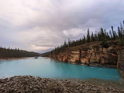 A photograph taken next to the shore of river. The river is a deep-blue colour. The shore is overed in rocks. The river banks are covered in pine trees.