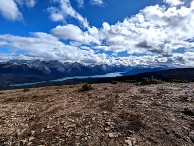 A photograph taken from a hiking trail, with grass and pine trees in the foreground, a distant lake can be seen, as well as snow-capped mountains in the background.