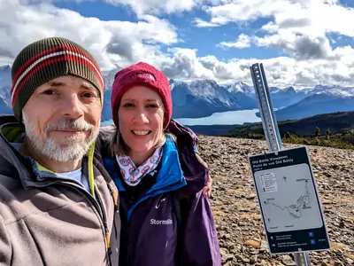 A photograph taken from a mountain peak, with a middle-aged male and female in the foreground standing next to a sign titled 'Old Baldy Viewpoint-2280 meters', in the distance is a lake, as well as snow-capped mountains.