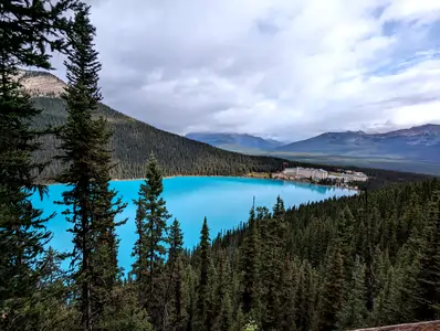 A photograph taken from the bank of a large deep-blue coloured lake. There's a large grand-looking hotel on the far shore. The lake and hotel are surrounded by pine forest.