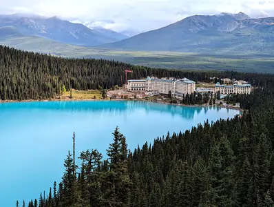 A photograph taken from the bank of a large deep-blue coloured lake. There's a large grand-looking hotel on the far shore. The lake and hotel are surrounded by pine forest.