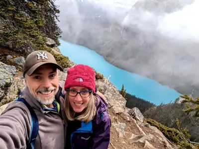 A photograph of a middle-aged male and female, who have their arms linked and are smiling at the camera. They are standing near the edge of a high cliff. In the background far below, is a deep-blue coloured lake.