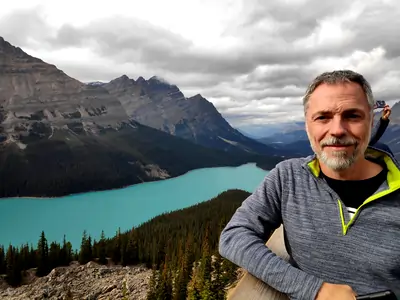 A photograph with a middle-aged man in the foreground, a deep-blue coloured blue lake in the middle ground, and mountains in the background.